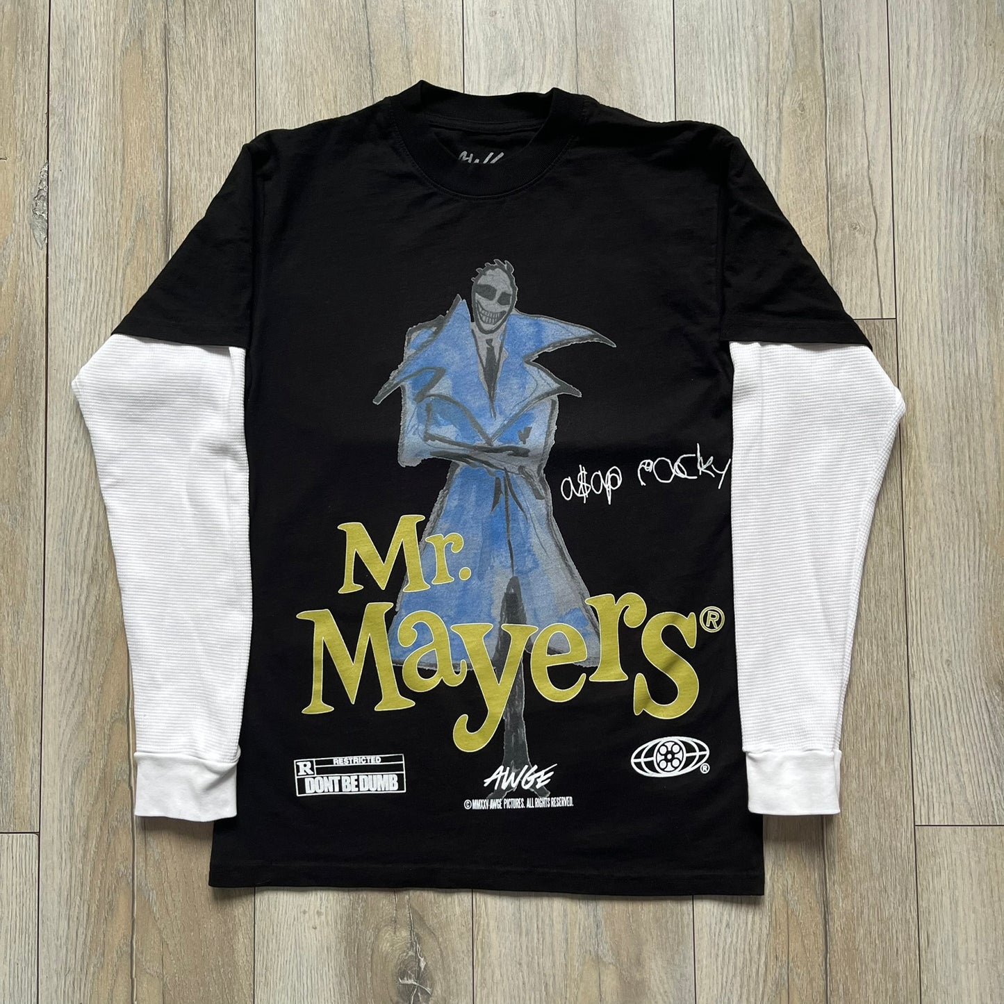 A$AP Rocky “Don’t Be Dumb” Mr Mayers Twofer Longsleeve is a limited release from the AWGE website tied to the rollout of Don’t Be Dumb by A$AP Rocky. The piece features a twofer construction with a black body and white sleeves, with a front graphic of the Mr Mayers figure in a blue trench coat with “Mr Mayers” text and small AWGE logos. The back includes a framed graphic with “Don’t Be Dumb” text, using a lenticular-style vinyl panel that shifts between the Mr Mayers figure and A$AP Rocky in the same outfit