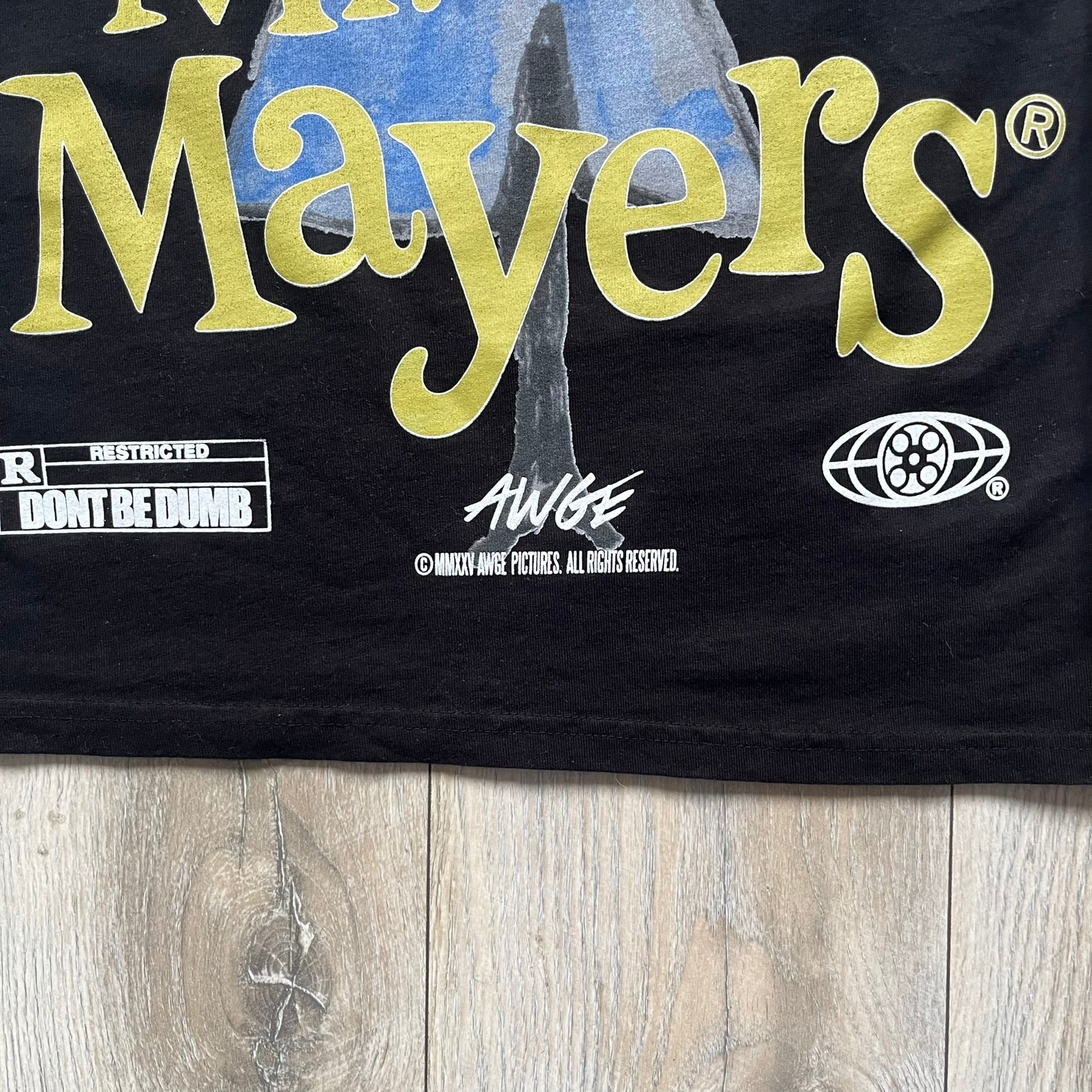 A$AP Rocky “Don’t Be Dumb” Mr Mayers Twofer Longsleeve is a limited release from the AWGE website tied to the rollout of Don’t Be Dumb by A$AP Rocky. The piece features a twofer construction with a black body and white sleeves, with a front graphic of the Mr Mayers figure in a blue trench coat with “Mr Mayers” text and small AWGE logos. The back includes a framed graphic with “Don’t Be Dumb” text, using a lenticular-style vinyl panel that shifts between the Mr Mayers figure and A$AP Rocky in the same outfit