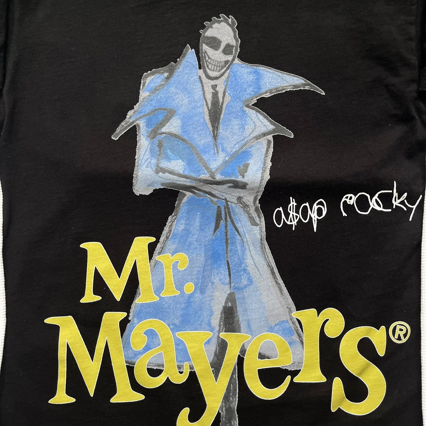A$AP Rocky “Don’t Be Dumb” Mr Mayers Twofer Longsleeve is a limited release from the AWGE website tied to the rollout of Don’t Be Dumb by A$AP Rocky. The piece features a twofer construction with a black body and white sleeves, with a front graphic of the Mr Mayers figure in a blue trench coat with “Mr Mayers” text and small AWGE logos. The back includes a framed graphic with “Don’t Be Dumb” text, using a lenticular-style vinyl panel that shifts between the Mr Mayers figure and A$AP Rocky in the same outfit