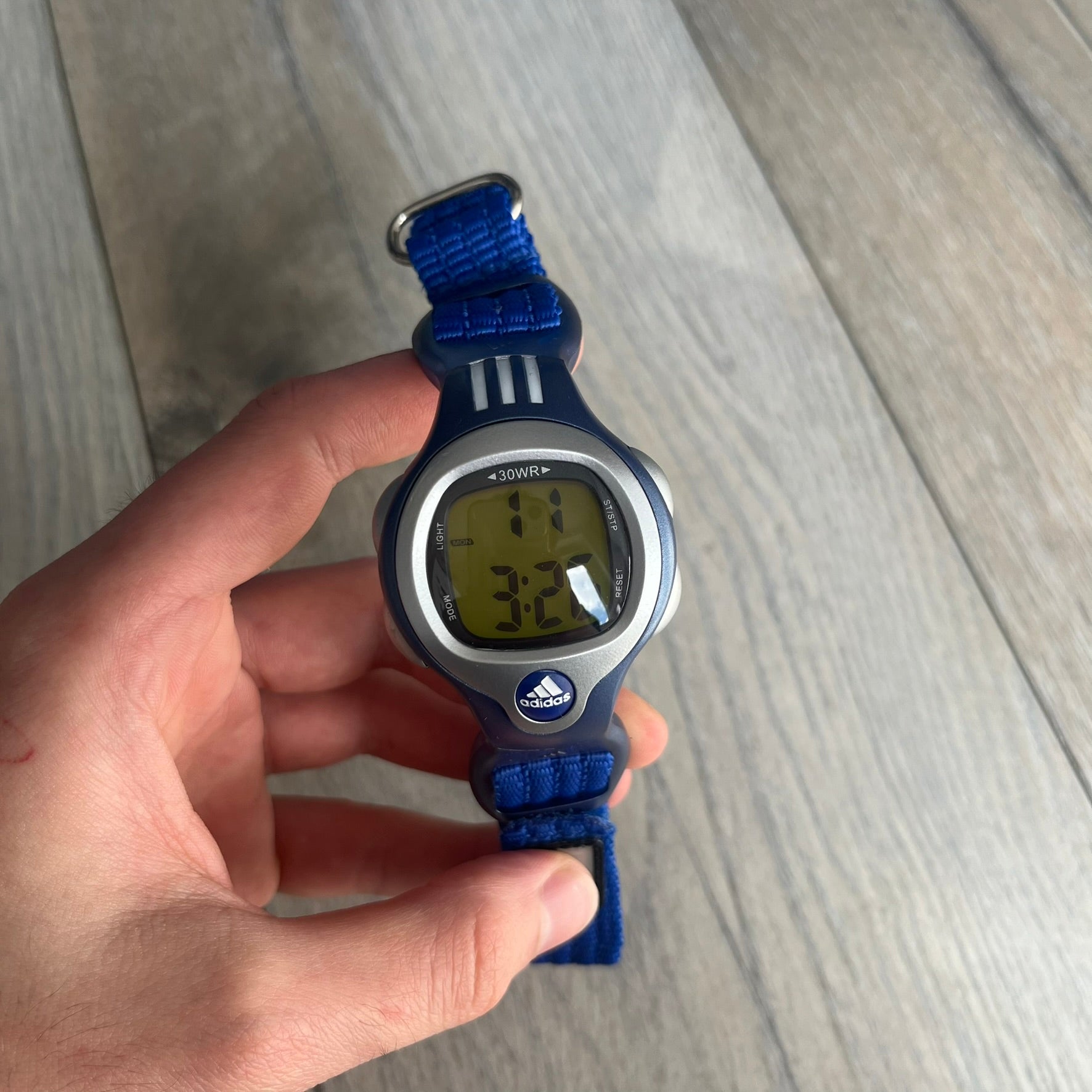Adidas 2000s Blue 10-0117 Digital Watch (model X-003) is a blue digital watch with a silver bezel and ergonomic case. It features a blue nylon Velcro strap, four side buttons, and a blue-green backlight display showing Adidas logo. The design includes three white stripes on the case and a silver Adidas logo button below the screen. The stainless steel back is engraved and marked water resistant up to 3 ATM, with an Adidas logo patch on the strap.