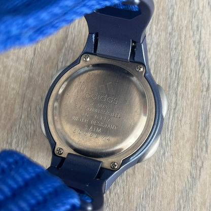 Adidas 2000s Blue 10-0117 Digital Watch (model X-003) is a blue digital watch with a silver bezel and ergonomic case. It features a blue nylon Velcro strap, four side buttons, and a blue-green backlight display showing Adidas logo. The design includes three white stripes on the case and a silver Adidas logo button below the screen. The stainless steel back is engraved and marked water resistant up to 3 ATM, with an Adidas logo patch on the strap.