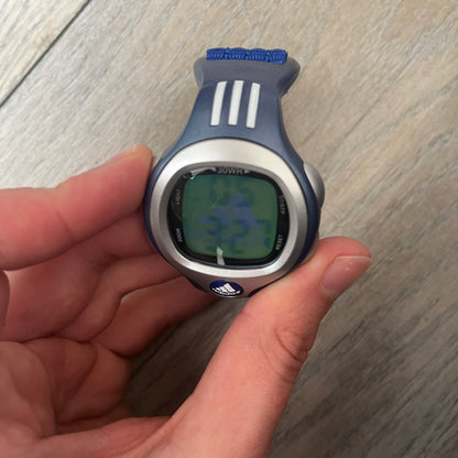 Adidas 2000s Blue 10-0117 Digital Watch (model X-003) is a blue digital watch with a silver bezel and ergonomic case. It features a blue nylon Velcro strap, four side buttons, and a blue-green backlight display showing Adidas logo. The design includes three white stripes on the case and a silver Adidas logo button below the screen. The stainless steel back is engraved and marked water resistant up to 3 ATM, with an Adidas logo patch on the strap.