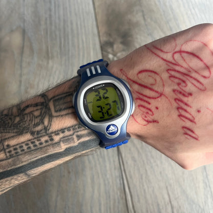 Adidas 2000s Blue 10-0117 Digital Watch (model X-003) is a blue digital watch with a silver bezel and ergonomic case. It features a blue nylon Velcro strap, four side buttons, and a blue-green backlight display showing Adidas logo. The design includes three white stripes on the case and a silver Adidas logo button below the screen. The stainless steel back is engraved and marked water resistant up to 3 ATM, with an Adidas logo patch on the strap.