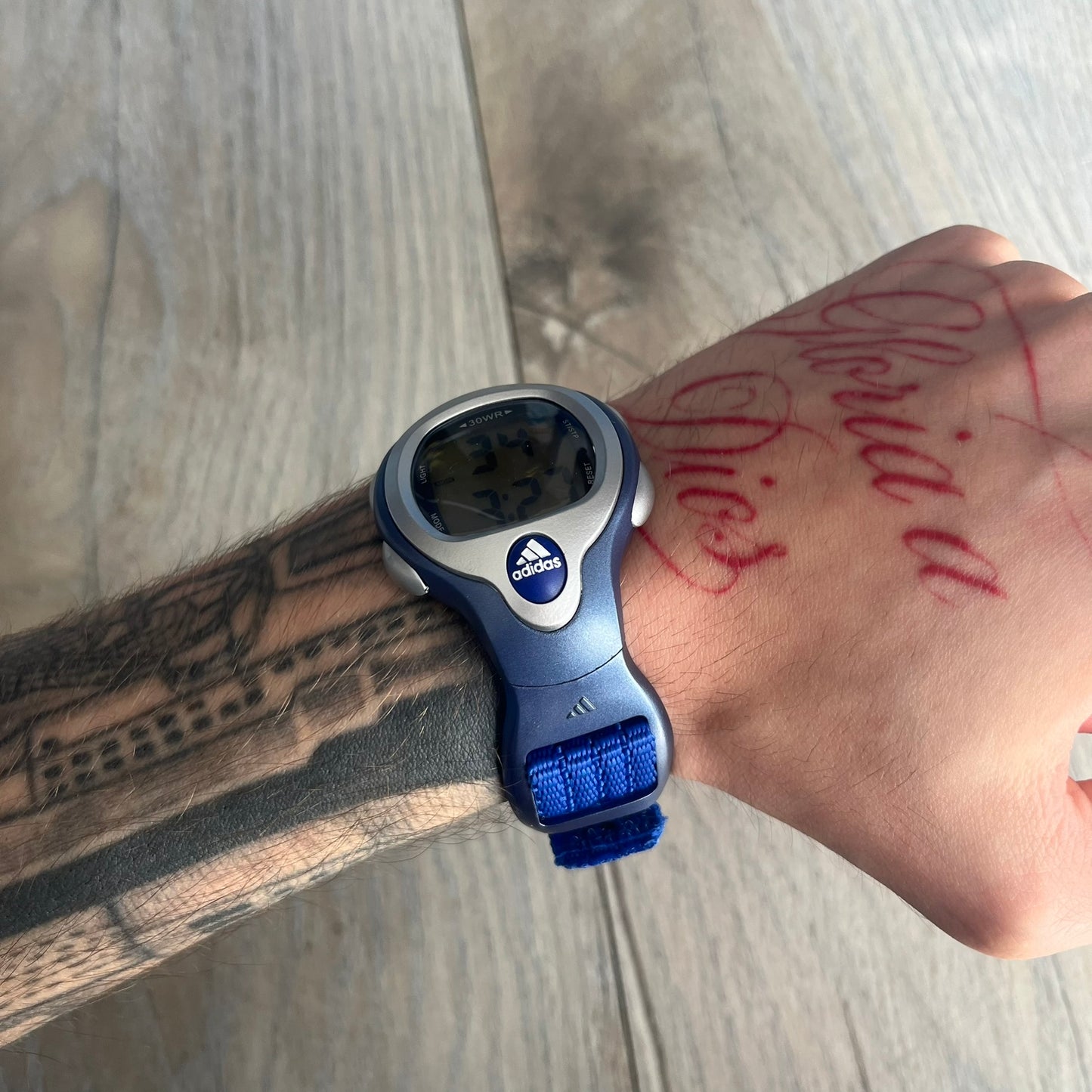 Adidas 2000s Blue 10-0117 Digital Watch (model X-003) is a blue digital watch with a silver bezel and ergonomic case. It features a blue nylon Velcro strap, four side buttons, and a blue-green backlight display showing Adidas logo. The design includes three white stripes on the case and a silver Adidas logo button below the screen. The stainless steel back is engraved and marked water resistant up to 3 ATM, with an Adidas logo patch on the strap.