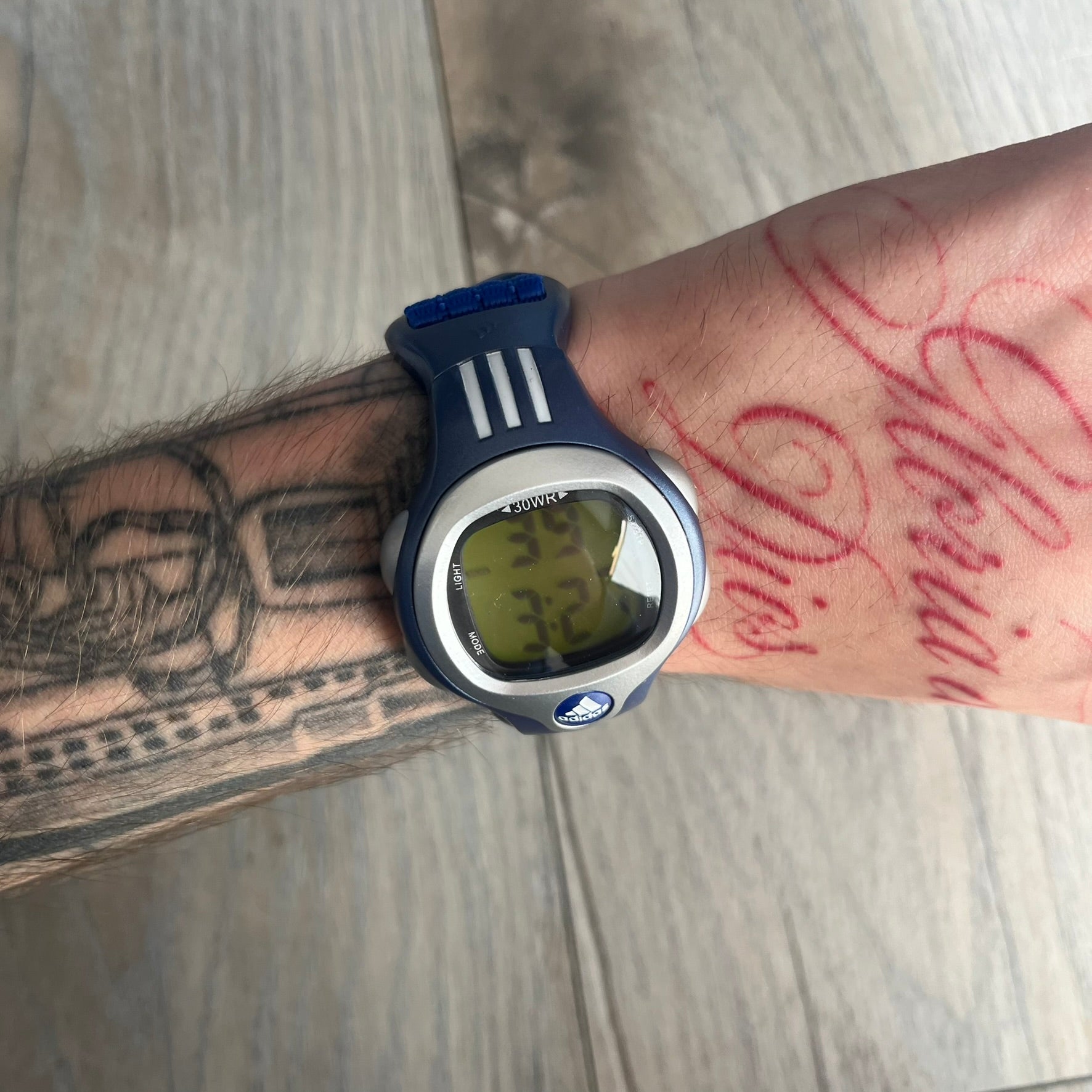 Adidas 2000s Blue 10-0117 Digital Watch (model X-003) is a blue digital watch with a silver bezel and ergonomic case. It features a blue nylon Velcro strap, four side buttons, and a blue-green backlight display showing Adidas logo. The design includes three white stripes on the case and a silver Adidas logo button below the screen. The stainless steel back is engraved and marked water resistant up to 3 ATM, with an Adidas logo patch on the strap.
