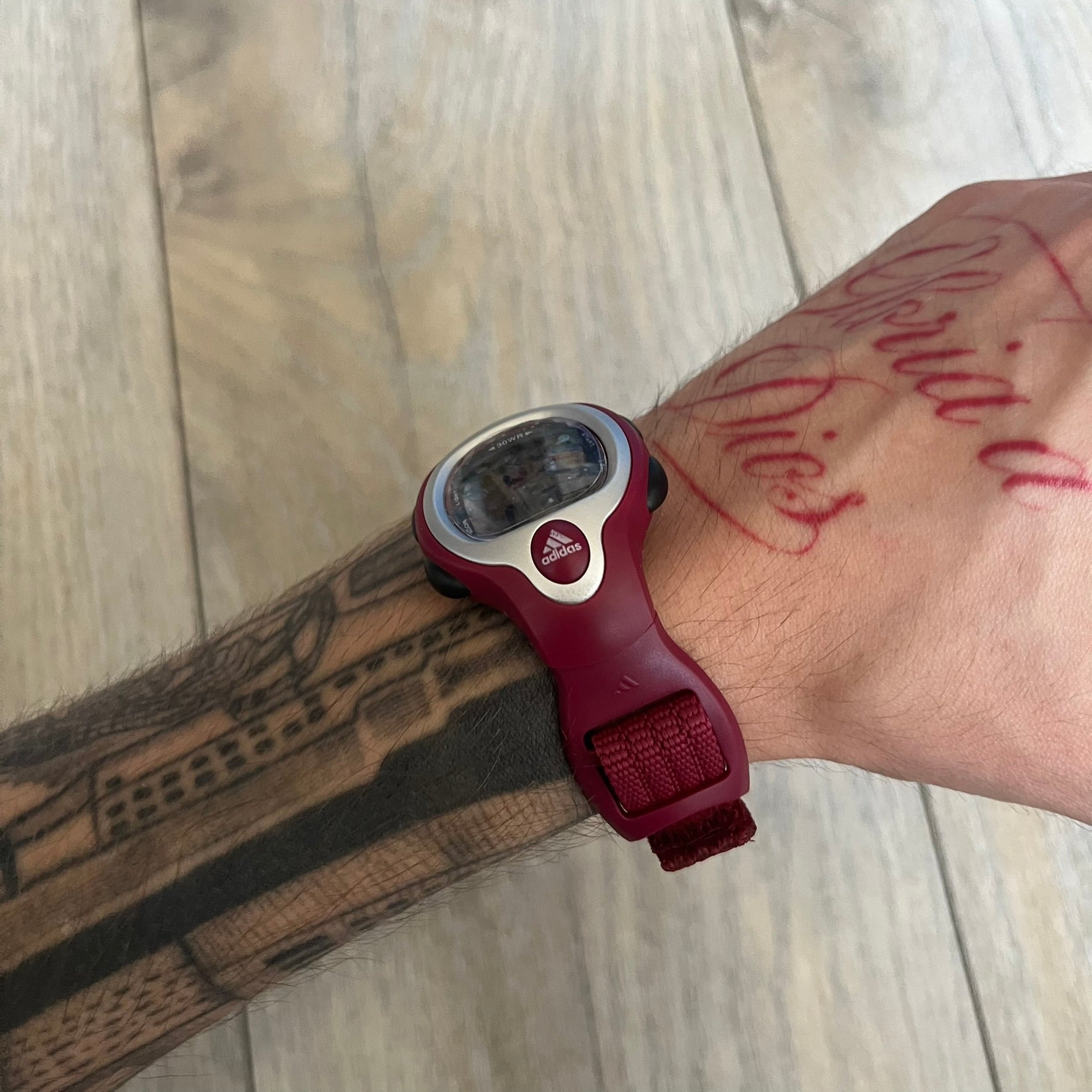 Adidas 2000s Burgundy 10-0117 Digital Watch (model X-003) is a burgundy digital watch with a silver bezel and ergonomic case. It features a burgundy nylon Velcro strap and four side buttons. The design includes three white stripes on the case and a silver Adidas logo button below the screen. The stainless steel back is engraved and marked water resistant up to 3 ATM, with an Adidas logo patch on the strap.