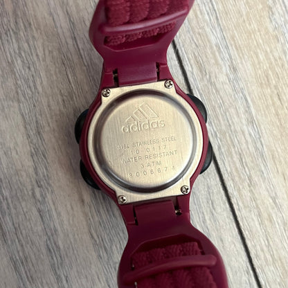 Adidas 2000s Burgundy 10-0117 Digital Watch (model X-003) is a burgundy digital watch with a silver bezel and ergonomic case. It features a burgundy nylon Velcro strap and four side buttons. The design includes three white stripes on the case and a silver Adidas logo button below the screen. The stainless steel back is engraved and marked water resistant up to 3 ATM, with an Adidas logo patch on the strap.