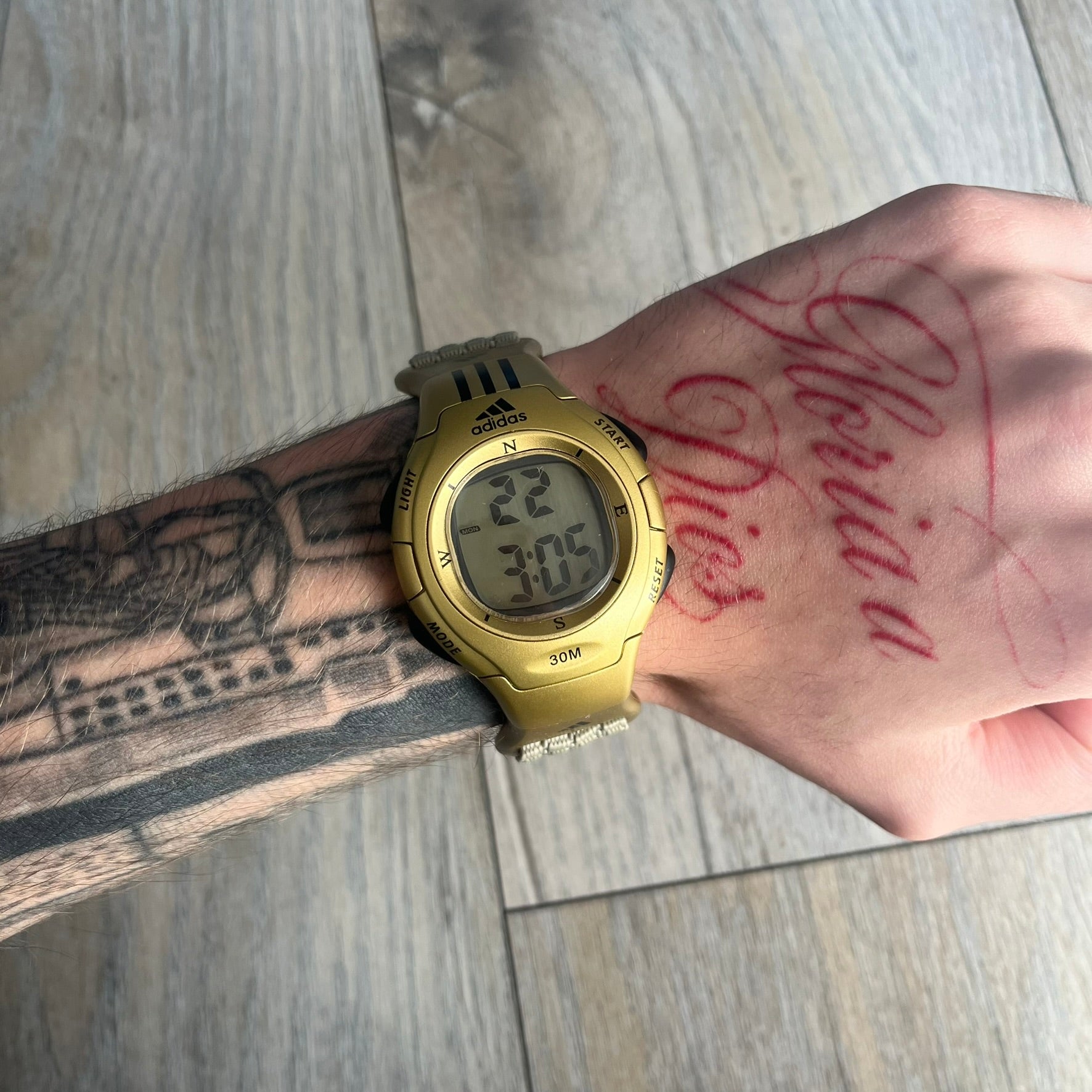 Adidas 2000s Gold 10-0117 Digital Watch (model X-003) is a fully gold digital watch with big and ergonomic case. It features a gold plastic and nylon strap, four side buttons, and a blue-green backlight display showing Adidas logo. The design includes three black stripes on the case and a black Adidas logo button above the screen. The stainless steel back is engraved and marked water resistant up to 3 ATM.