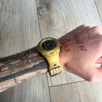 Adidas 2000s Gold 10-0117 Digital Watch (model X-003) is a fully gold digital watch with big and ergonomic case. It features a gold plastic and nylon strap, four side buttons, and a blue-green backlight display showing Adidas logo. The design includes three black stripes on the case and a black Adidas logo button above the screen. The stainless steel back is engraved and marked water resistant up to 3 ATM.