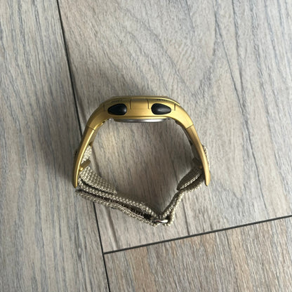 Adidas 2000s Gold 10-0117 Digital Watch (model X-003) is a fully gold digital watch with big and ergonomic case. It features a gold plastic and nylon strap, four side buttons, and a blue-green backlight display showing Adidas logo. The design includes three black stripes on the case and a black Adidas logo button above the screen. The stainless steel back is engraved and marked water resistant up to 3 ATM.