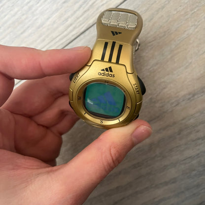 Adidas 2000s Gold 10-0117 Digital Watch (model X-003) is a fully gold digital watch with big and ergonomic case. It features a gold plastic and nylon strap, four side buttons, and a blue-green backlight display showing Adidas logo. The design includes three black stripes on the case and a black Adidas logo button above the screen. The stainless steel back is engraved and marked water resistant up to 3 ATM.