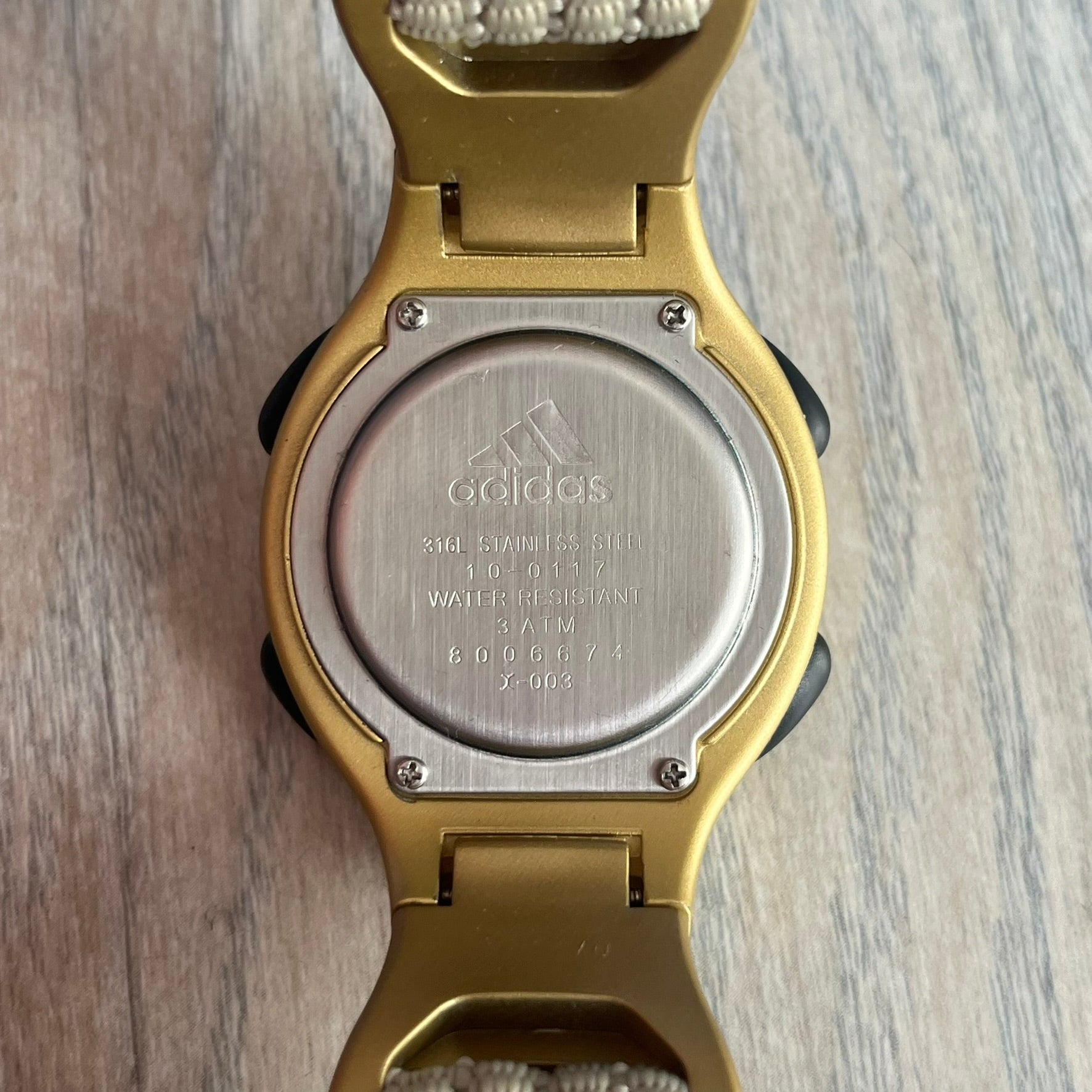 Adidas 2000s Gold 10-0117 Digital Watch (model X-003) is a fully gold digital watch with big and ergonomic case. It features a gold plastic and nylon strap, four side buttons, and a blue-green backlight display showing Adidas logo. The design includes three black stripes on the case and a black Adidas logo button above the screen. The stainless steel back is engraved and marked water resistant up to 3 ATM.
