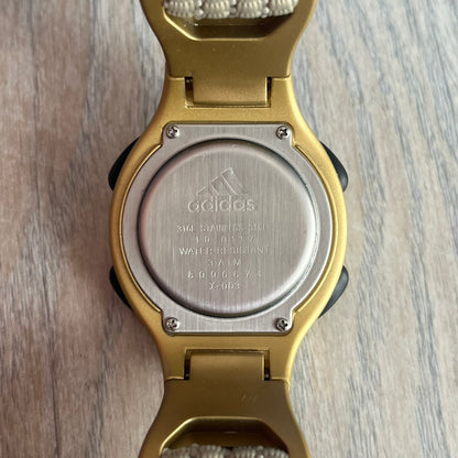 Adidas 2000s Gold 10-0117 Digital Watch (model X-003) is a fully gold digital watch with big and ergonomic case. It features a gold plastic and nylon strap, four side buttons, and a blue-green backlight display showing Adidas logo. The design includes three black stripes on the case and a black Adidas logo button above the screen. The stainless steel back is engraved and marked water resistant up to 3 ATM.