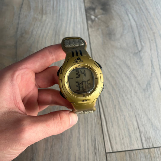Adidas 2000s Gold 10-0117 Digital Watch (model X-003) is a fully gold digital watch with big and ergonomic case. It features a gold plastic and nylon strap, four side buttons, and a blue-green backlight display showing Adidas logo. The design includes three black stripes on the case and a black Adidas logo button above the screen. The stainless steel back is engraved and marked water resistant up to 3 ATM.