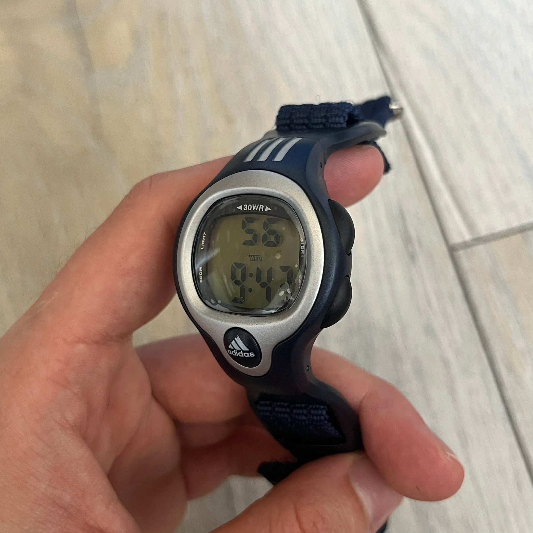 Adidas 2000s Navy 10-0117 Digital Watch (model X-003) is a navy digital watch with a silver bezel and ergonomic case. It features a navy nylon Velcro strap and four side buttons. The design includes three white stripes on the case and a silver Adidas logo button below the screen. The stainless steel back is engraved and marked water resistant up to 3 ATM, with an Adidas logo patch on the strap.