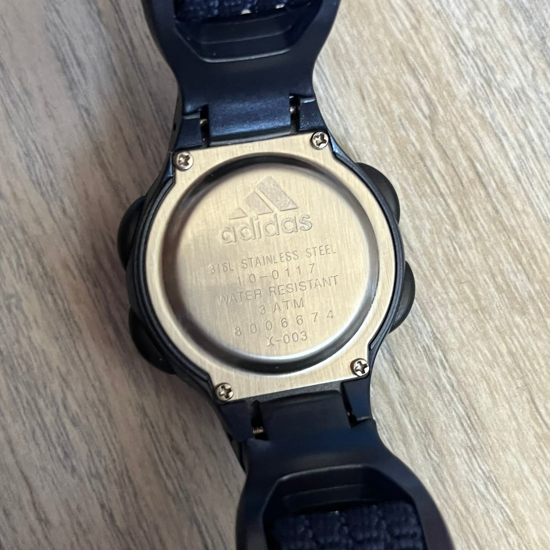 Adidas 2000s Navy 10-0117 Digital Watch (model X-003) is a navy digital watch with a silver bezel and ergonomic case. It features a navy nylon Velcro strap and four side buttons. The design includes three white stripes on the case and a silver Adidas logo button below the screen. The stainless steel back is engraved and marked water resistant up to 3 ATM, with an Adidas logo patch on the strap.