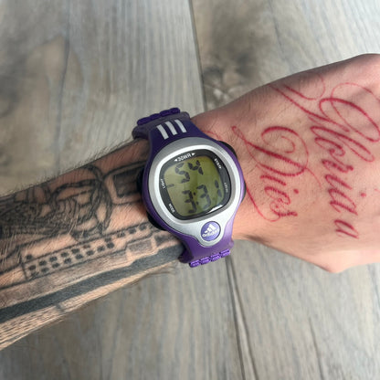 Adidas 2000s Purple 10-0117 Digital Watch (model X-003) is a purple digital watch with a silver bezel and ergonomic case. It features a purple nylon Velcro strap, four side buttons, and a blue-green backlight display showing Adidas logo. The design includes three white stripes on the case and a silver Adidas logo button below the screen. The stainless steel back is engraved and marked water resistant up to 3 ATM, with an Adidas logo patch on the strap
