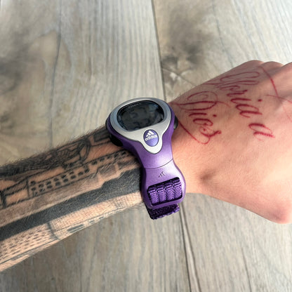 Adidas 2000s Purple 10-0117 Digital Watch (model X-003) is a purple digital watch with a silver bezel and ergonomic case. It features a purple nylon Velcro strap, four side buttons, and a blue-green backlight display showing Adidas logo. The design includes three white stripes on the case and a silver Adidas logo button below the screen. The stainless steel back is engraved and marked water resistant up to 3 ATM, with an Adidas logo patch on the strap