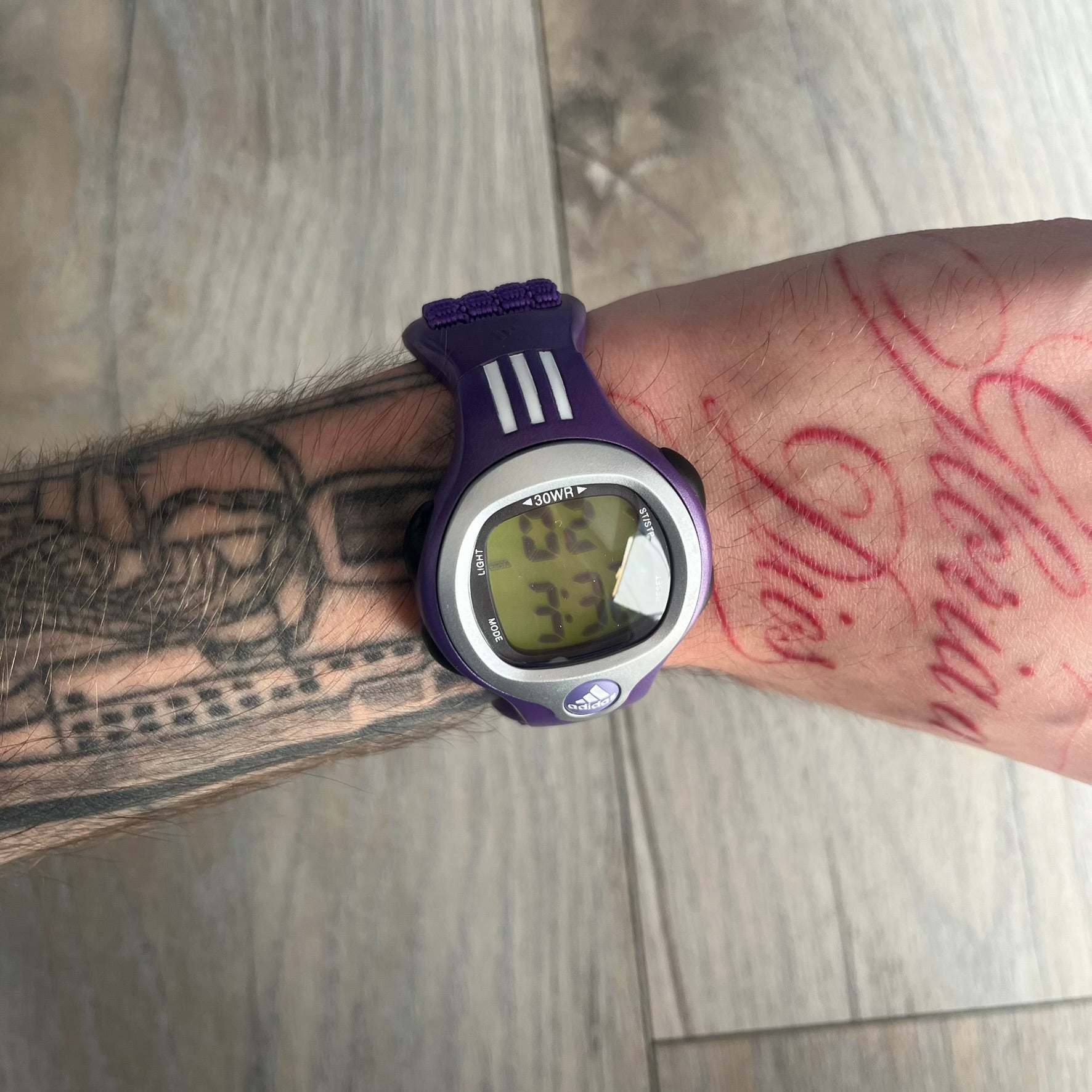 Adidas 2000s Purple 10-0117 Digital Watch (model X-003) is a purple digital watch with a silver bezel and ergonomic case. It features a purple nylon Velcro strap, four side buttons, and a blue-green backlight display showing Adidas logo. The design includes three white stripes on the case and a silver Adidas logo button below the screen. The stainless steel back is engraved and marked water resistant up to 3 ATM, with an Adidas logo patch on the strap