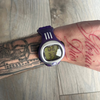 Adidas 2000s Purple 10-0117 Digital Watch (model X-003) is a purple digital watch with a silver bezel and ergonomic case. It features a purple nylon Velcro strap, four side buttons, and a blue-green backlight display showing Adidas logo. The design includes three white stripes on the case and a silver Adidas logo button below the screen. The stainless steel back is engraved and marked water resistant up to 3 ATM, with an Adidas logo patch on the strap