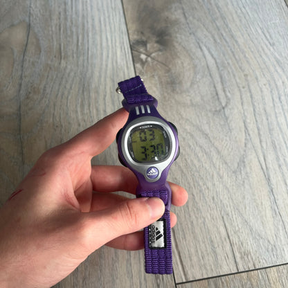 Adidas 2000s Purple 10-0117 Digital Watch (model X-003) is a purple digital watch with a silver bezel and ergonomic case. It features a purple nylon Velcro strap, four side buttons, and a blue-green backlight display showing Adidas logo. The design includes three white stripes on the case and a silver Adidas logo button below the screen. The stainless steel back is engraved and marked water resistant up to 3 ATM, with an Adidas logo patch on the strap