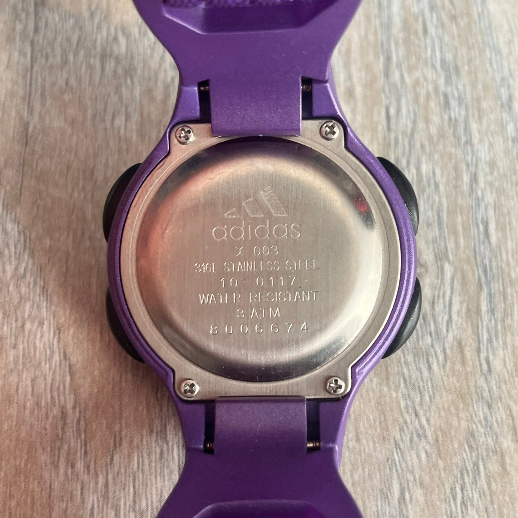 Adidas 2000s Purple 10-0117 Digital Watch (model X-003) is a purple digital watch with a silver bezel and ergonomic case. It features a purple nylon Velcro strap, four side buttons, and a blue-green backlight display showing Adidas logo. The design includes three white stripes on the case and a silver Adidas logo button below the screen. The stainless steel back is engraved and marked water resistant up to 3 ATM, with an Adidas logo patch on the strap