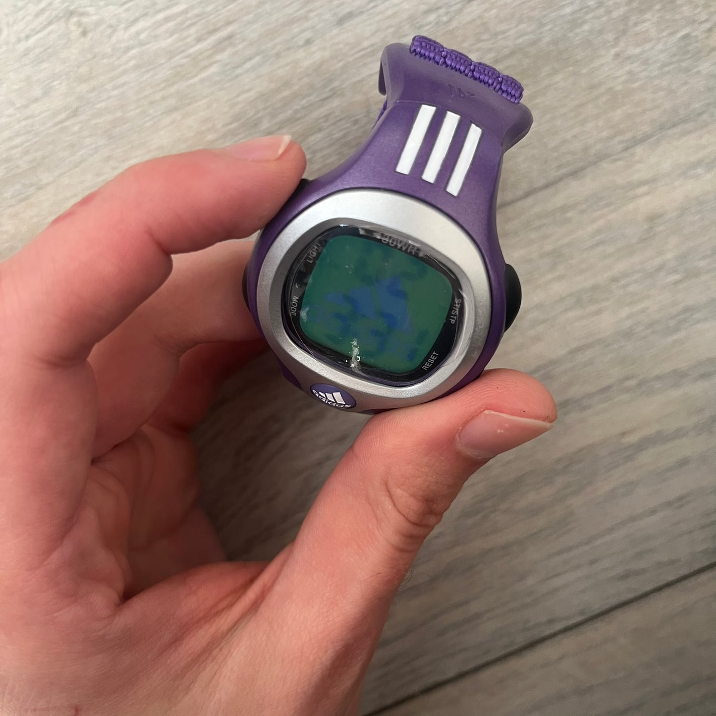 Adidas 2000s Purple 10-0117 Digital Watch (model X-003) is a purple digital watch with a silver bezel and ergonomic case. It features a purple nylon Velcro strap, four side buttons, and a blue-green backlight display showing Adidas logo. The design includes three white stripes on the case and a silver Adidas logo button below the screen. The stainless steel back is engraved and marked water resistant up to 3 ATM, with an Adidas logo patch on the strap
