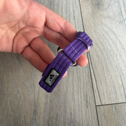 Adidas 2000s Purple 10-0117 Digital Watch (model X-003) is a purple digital watch with a silver bezel and ergonomic case. It features a purple nylon Velcro strap, four side buttons, and a blue-green backlight display showing Adidas logo. The design includes three white stripes on the case and a silver Adidas logo button below the screen. The stainless steel back is engraved and marked water resistant up to 3 ATM, with an Adidas logo patch on the strap