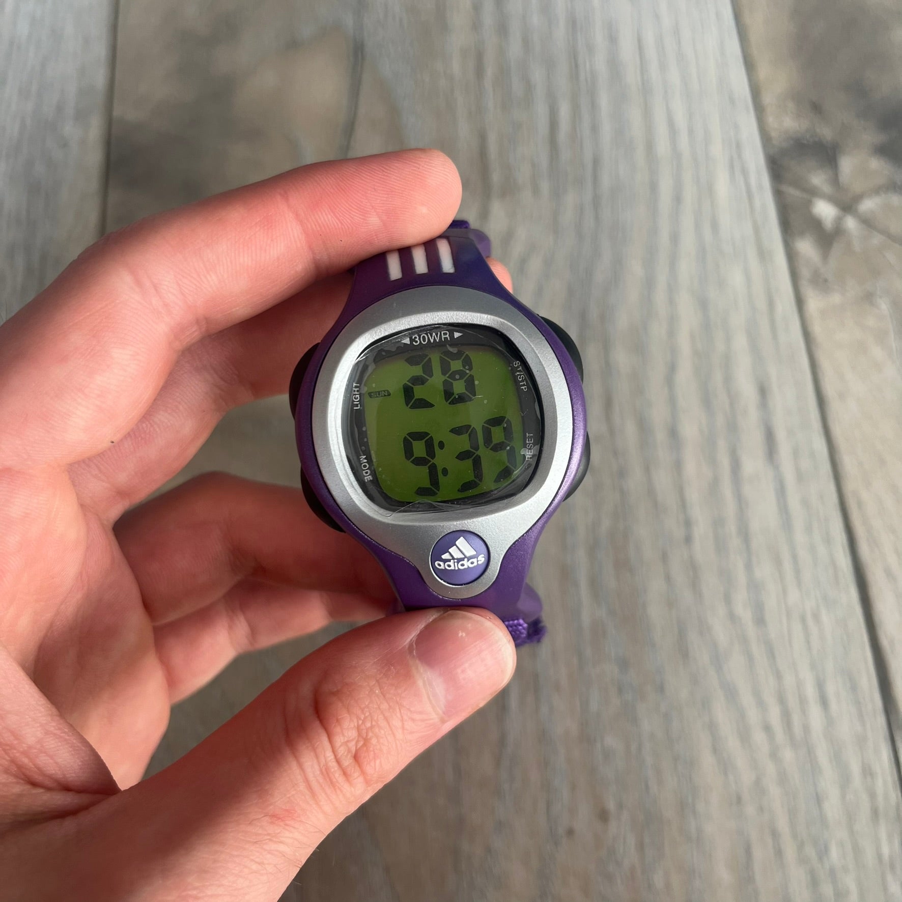 Adidas 2000s Purple 10-0117 Digital Watch (model X-003) is a purple digital watch with a silver bezel and ergonomic case. It features a purple nylon Velcro strap, four side buttons, and a blue-green backlight display showing Adidas logo. The design includes three white stripes on the case and a silver Adidas logo button below the screen. The stainless steel back is engraved and marked water resistant up to 3 ATM, with an Adidas logo patch on the strap.