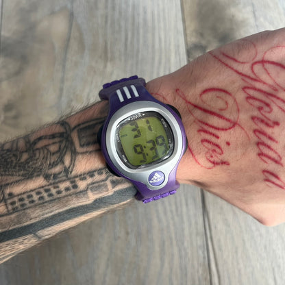 Adidas 2000s Purple 10-0117 Digital Watch (model X-003) is a purple digital watch with a silver bezel and ergonomic case. It features a purple nylon Velcro strap, four side buttons, and a blue-green backlight display showing Adidas logo. The design includes three white stripes on the case and a silver Adidas logo button below the screen. The stainless steel back is engraved and marked water resistant up to 3 ATM, with an Adidas logo patch on the strap.