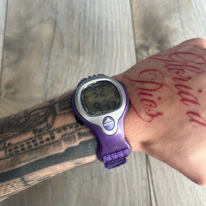 Adidas 2000s Purple 10-0117 Digital Watch (model X-003) is a purple digital watch with a silver bezel and ergonomic case. It features a purple nylon Velcro strap, four side buttons, and a blue-green backlight display showing Adidas logo. The design includes three white stripes on the case and a silver Adidas logo button below the screen. The stainless steel back is engraved and marked water resistant up to 3 ATM, with an Adidas logo patch on the strap.