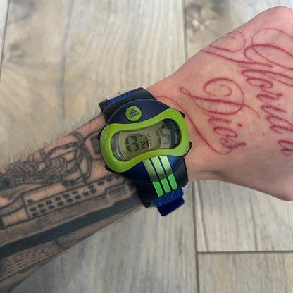 Adidas 2000’s Sports 10-0099L-003 Digital Watch is a digital wristwatch featuring a PC and fibre glass case with a 316L stainless steel caseback. The watch has a green, blue, and black color scheme with a blue backlight display and is water resistant to 100 meters. It includes a Velcro strap with clip closure for adjustable fit and is marked with reference number 10-0099L-003 on the back. The watch comes with an adidas box.