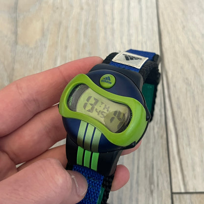 Adidas 2000’s Sports 10-0099L-003 Digital Watch is a digital wristwatch featuring a PC and fibre glass case with a 316L stainless steel caseback. The watch has a green, blue, and black color scheme with a blue backlight display and is water resistant to 100 meters. It includes a Velcro strap with clip closure for adjustable fit and is marked with reference number 10-0099L-003 on the back. The watch comes with an adidas box.
