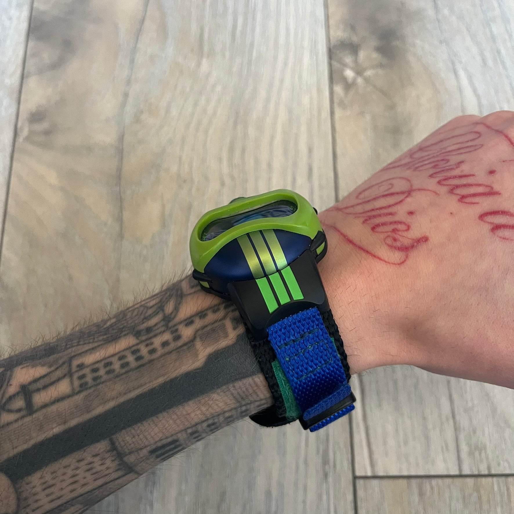 Adidas 2000’s Sports 10-0099L-003 Digital Watch is a digital wristwatch featuring a PC and fibre glass case with a 316L stainless steel caseback. The watch has a green, blue, and black color scheme with a blue backlight display and is water resistant to 100 meters. It includes a Velcro strap with clip closure for adjustable fit and is marked with reference number 10-0099L-003 on the back. The watch comes with an adidas box.