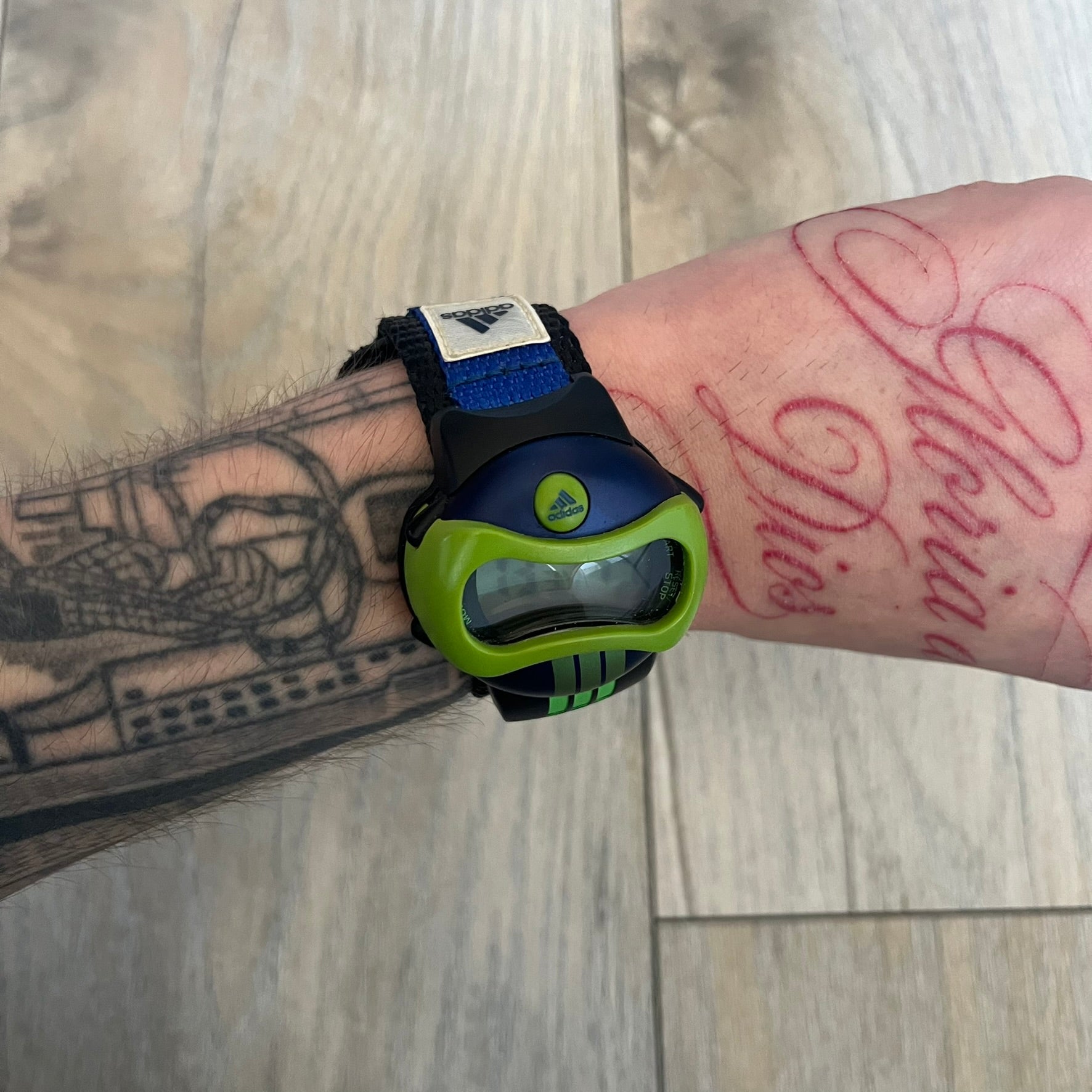 Adidas 2000’s Sports 10-0099L-003 Digital Watch is a digital wristwatch featuring a PC and fibre glass case with a 316L stainless steel caseback. The watch has a green, blue, and black color scheme with a blue backlight display and is water resistant to 100 meters. It includes a Velcro strap with clip closure for adjustable fit and is marked with reference number 10-0099L-003 on the back. The watch comes with an adidas box.