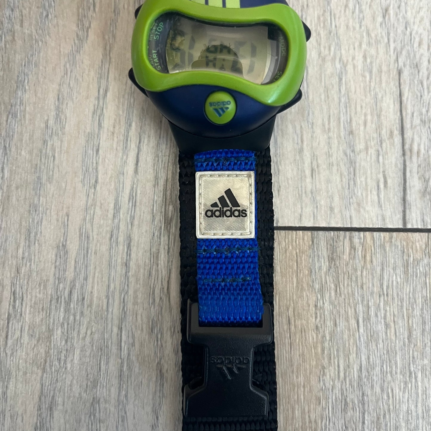 Adidas 2000’s Sports 10-0099L-003 Digital Watch is a digital wristwatch featuring a PC and fibre glass case with a 316L stainless steel caseback. The watch has a green, blue, and black color scheme with a blue backlight display and is water resistant to 100 meters. It includes a Velcro strap with clip closure for adjustable fit and is marked with reference number 10-0099L-003 on the back. The watch comes with an adidas box.