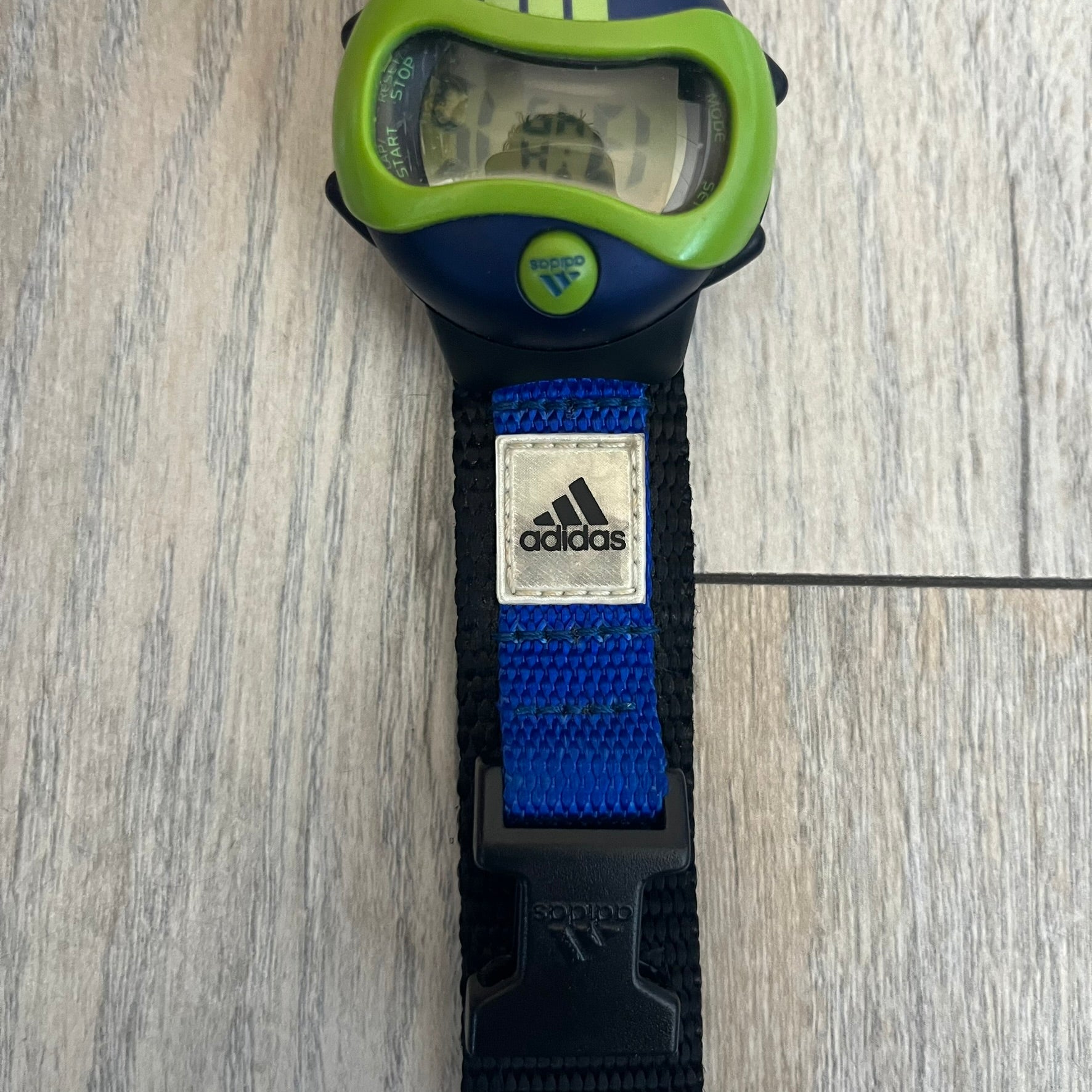 Adidas 2000’s Sports 10-0099L-003 Digital Watch is a digital wristwatch featuring a PC and fibre glass case with a 316L stainless steel caseback. The watch has a green, blue, and black color scheme with a blue backlight display and is water resistant to 100 meters. It includes a Velcro strap with clip closure for adjustable fit and is marked with reference number 10-0099L-003 on the back. The watch comes with an adidas box.