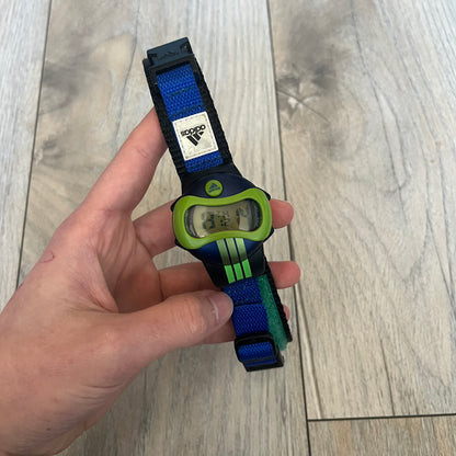 Adidas 2000’s Sports 10-0099L-003 Digital Watch is a digital wristwatch featuring a PC and fibre glass case with a 316L stainless steel caseback. The watch has a green, blue, and black color scheme with a blue backlight display and is water resistant to 100 meters. It includes a Velcro strap with clip closure for adjustable fit and is marked with reference number 10-0099L-003 on the back. The watch comes with an adidas box.