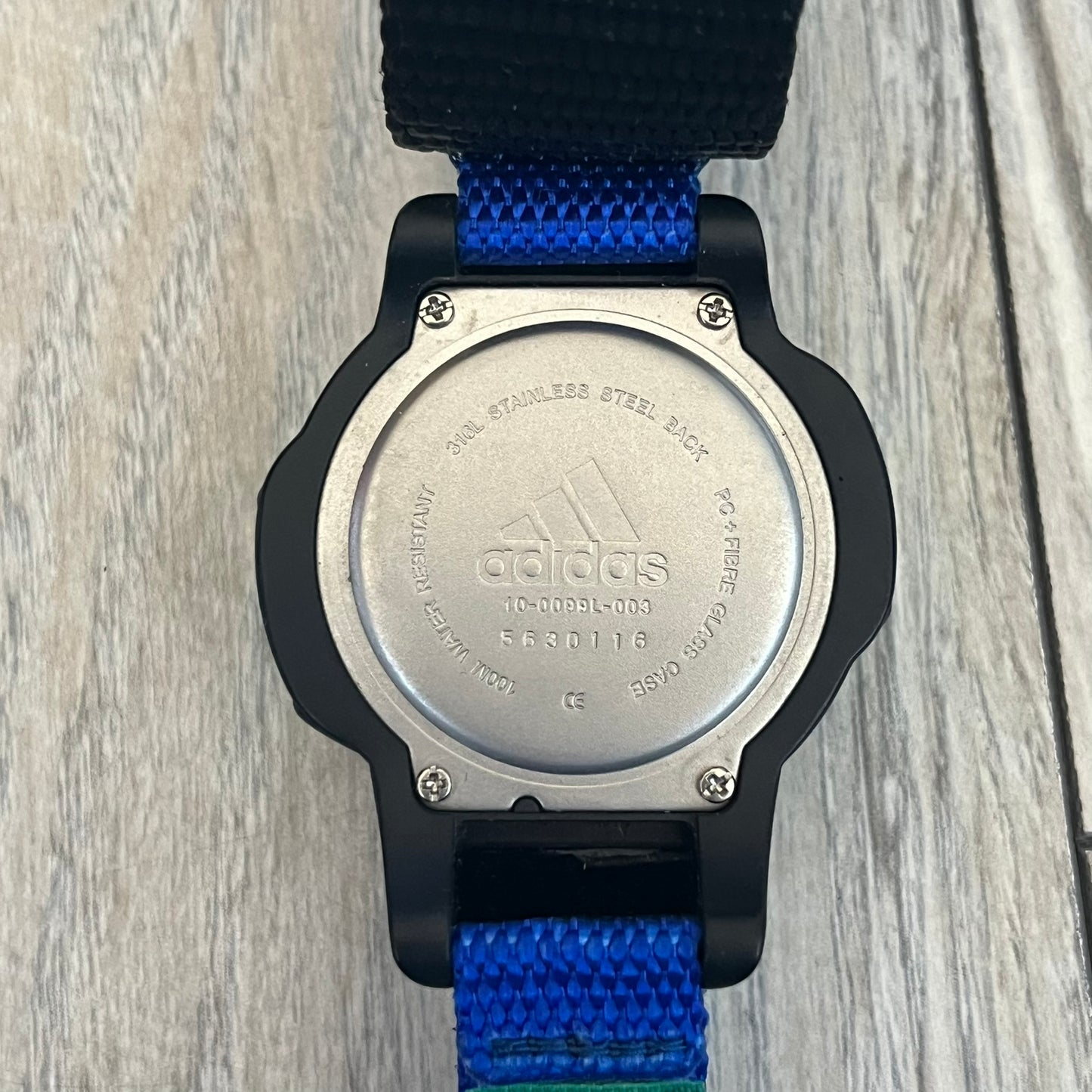 Adidas 2000’s Sports 10-0099L-003 Digital Watch is a digital wristwatch featuring a PC and fibre glass case with a 316L stainless steel caseback. The watch has a green, blue, and black color scheme with a blue backlight display and is water resistant to 100 meters. It includes a Velcro strap with clip closure for adjustable fit and is marked with reference number 10-0099L-003 on the back. The watch comes with an adidas box.