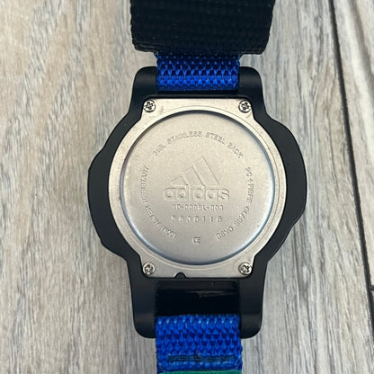 Adidas 2000’s Sports 10-0099L-003 Digital Watch is a digital wristwatch featuring a PC and fibre glass case with a 316L stainless steel caseback. The watch has a green, blue, and black color scheme with a blue backlight display and is water resistant to 100 meters. It includes a Velcro strap with clip closure for adjustable fit and is marked with reference number 10-0099L-003 on the back. The watch comes with an adidas box.