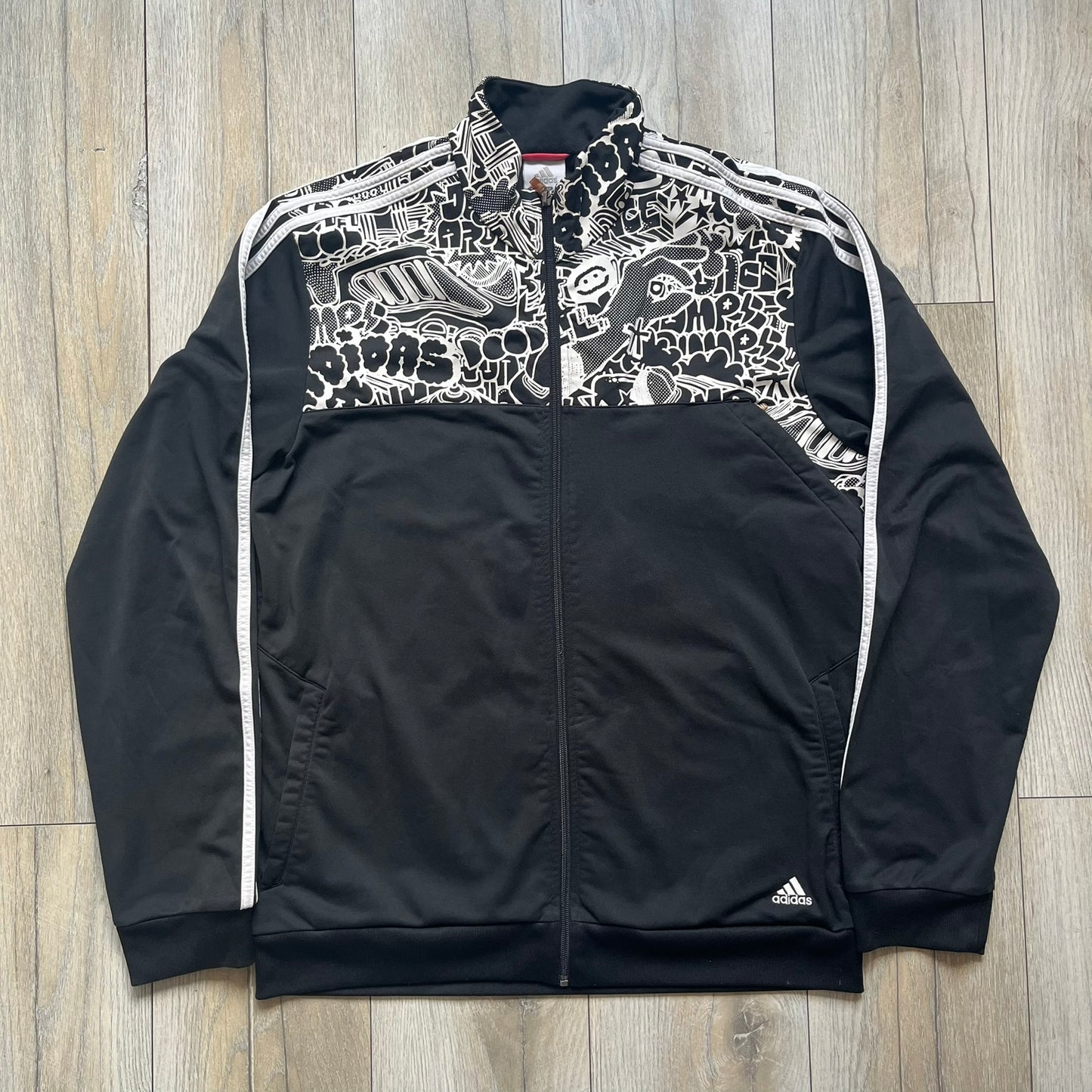 Adidas 2009 Doodle Track Jacket is a black track jacket with white Adidas stripes running down the sleeves. The upper chest area on both the front and back is covered in an all-over white doodle and graffiti-style print. It features a full zip closure and a small pocket on the left chest.

