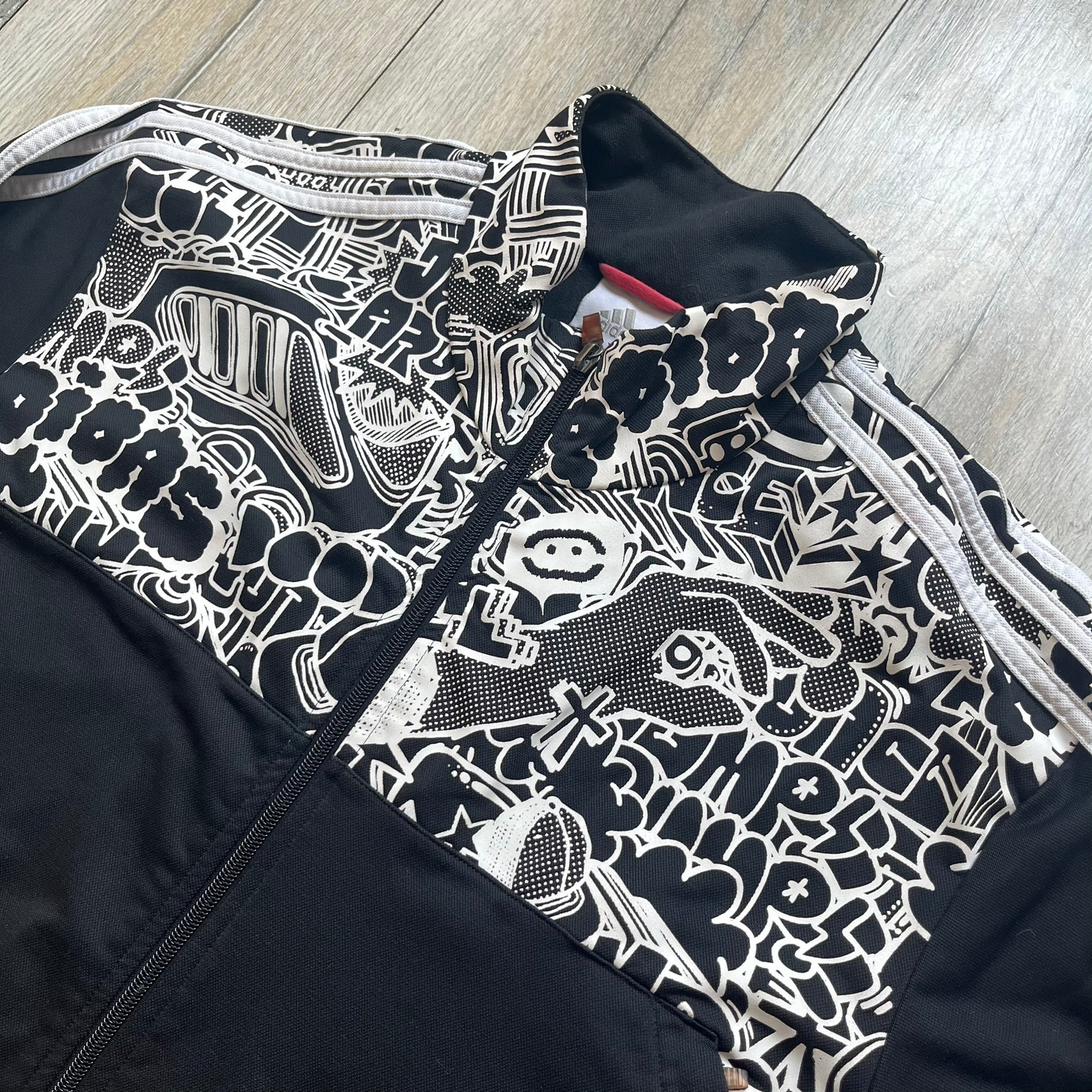 Adidas 2009 Doodle Track Jacket is a black track jacket with white Adidas stripes running down the sleeves. The upper chest area on both the front and back is covered in an all-over white doodle and graffiti-style print. It features a full zip closure and a small pocket on the left chest.
