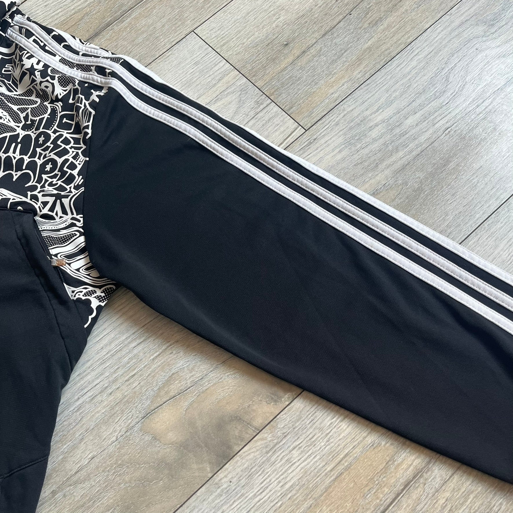 Adidas 2009 Doodle Track Jacket is a black track jacket with white Adidas stripes running down the sleeves. The upper chest area on both the front and back is covered in an all-over white doodle and graffiti-style print. It features a full zip closure and a small pocket on the left chest.
