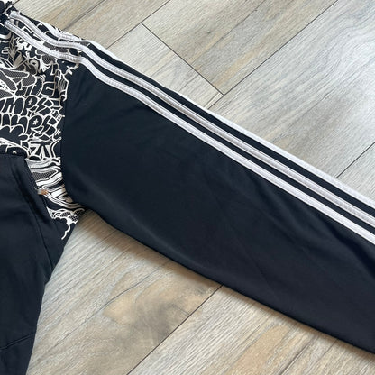 Adidas 2009 Doodle Track Jacket is a black track jacket with white Adidas stripes running down the sleeves. The upper chest area on both the front and back is covered in an all-over white doodle and graffiti-style print. It features a full zip closure and a small pocket on the left chest.
