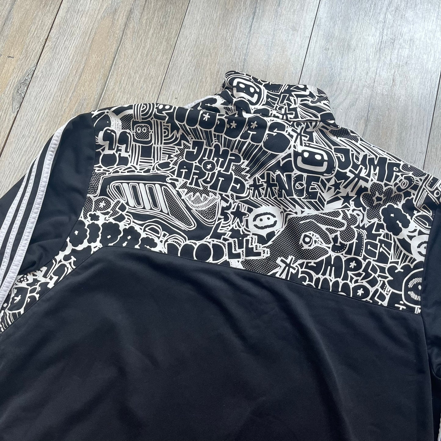Adidas 2009 Doodle Track Jacket is a black track jacket with white Adidas stripes running down the sleeves. The upper chest area on both the front and back is covered in an all-over white doodle and graffiti-style print. It features a full zip closure and a small pocket on the left chest.
