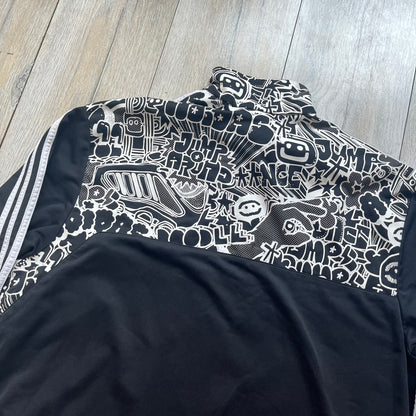 Adidas 2009 Doodle Track Jacket is a black track jacket with white Adidas stripes running down the sleeves. The upper chest area on both the front and back is covered in an all-over white doodle and graffiti-style print. It features a full zip closure and a small pocket on the left chest.
