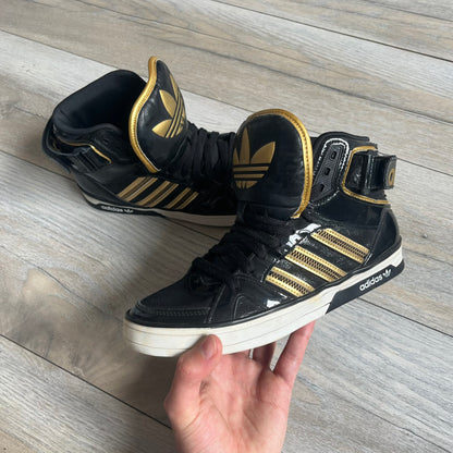 Adidas Space Diver Zip Pack Sneakers were released on November 18, 2012. The shoes feature a black upper with a glossy leather-like finish, gold detailing, and a white outsole and insole. The tongue has a large gold Trefoil logo with a gold outline, while the heel strap is printed with gold Adidas text. The sides feature three gold stripes designed as decorative zipper elements. A small white Adidas logo is also present on the upper.