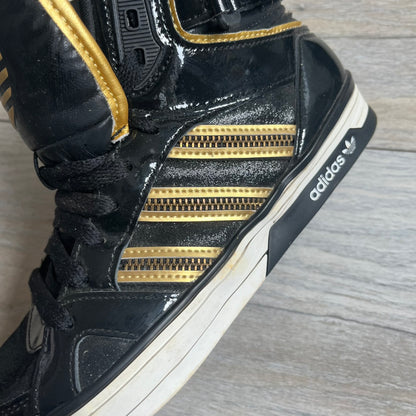 Adidas Space Diver Zip Pack Sneakers were released on November 18, 2012. The shoes feature a black upper with a glossy leather-like finish, gold detailing, and a white outsole and insole. The tongue has a large gold Trefoil logo with a gold outline, while the heel strap is printed with gold Adidas text. The sides feature three gold stripes designed as decorative zipper elements. A small white Adidas logo is also present on the upper.