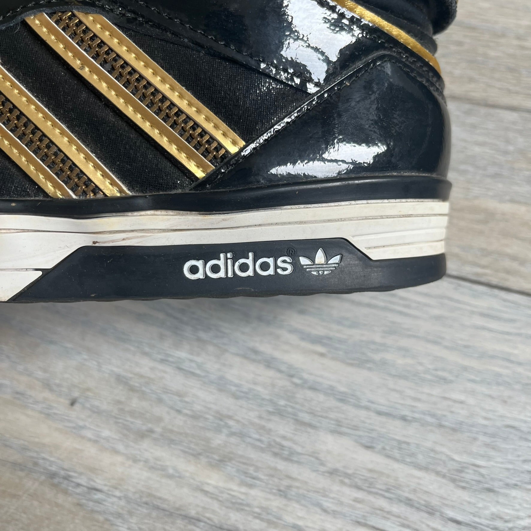 Adidas Space Diver Zip Pack Sneakers were released on November 18, 2012. The shoes feature a black upper with a glossy leather-like finish, gold detailing, and a white outsole and insole. The tongue has a large gold Trefoil logo with a gold outline, while the heel strap is printed with gold Adidas text. The sides feature three gold stripes designed as decorative zipper elements. A small white Adidas logo is also present on the upper.