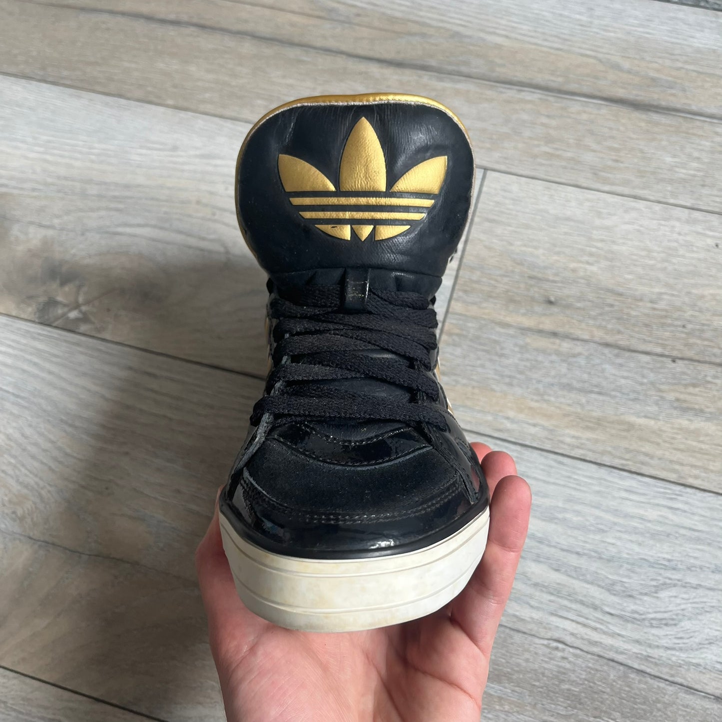 Adidas Space Diver Zip Pack Sneakers were released on November 18, 2012. The shoes feature a black upper with a glossy leather-like finish, gold detailing, and a white outsole and insole. The tongue has a large gold Trefoil logo with a gold outline, while the heel strap is printed with gold Adidas text. The sides feature three gold stripes designed as decorative zipper elements. A small white Adidas logo is also present on the upper.